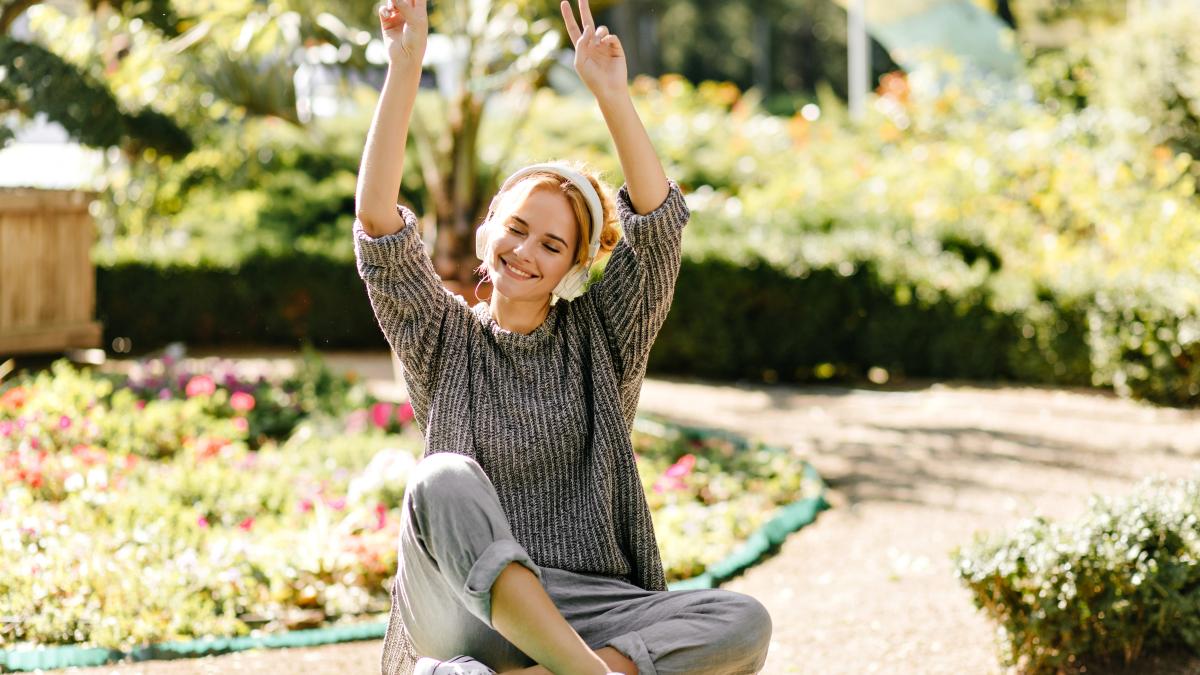 beautiful-woman-gray-oversized-sweater-is-smiling-listening-music-headphones-showing-peace-sign-background-garden_12150300